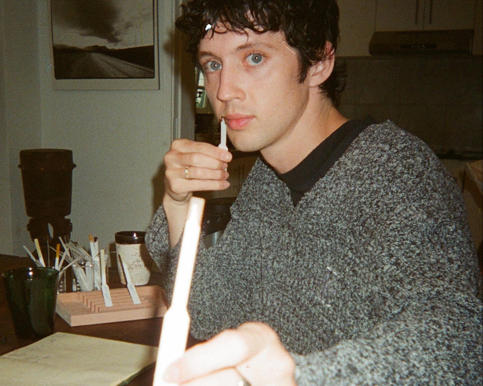 A man in a gray knit sweater holds a white paper near his mouth while sitting at a desk testing fragrance.