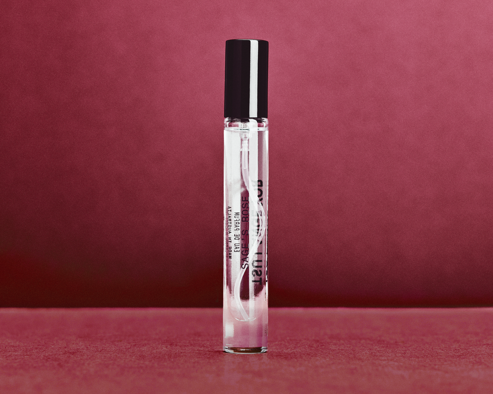 A clear cylindrical roller bottle with a black cap, standing upright against a textured dark red background. The bottle contains clear liquid and has black text printed on the side.